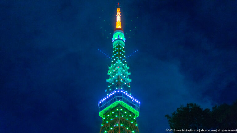 Upper Tokyo Tower glowing green, blue, and orange against a cloudy night sky.