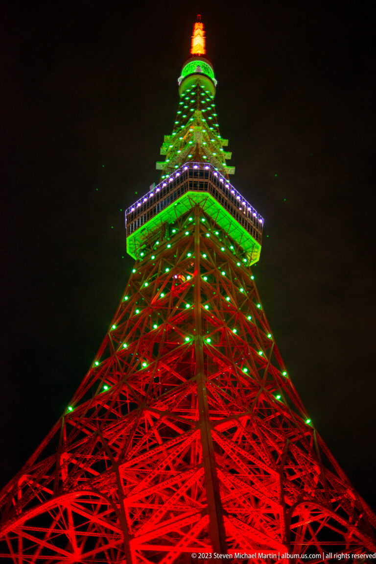 Low-angle view up Tokyo Tower’s red lattice toward green and blue-lit upper levels.