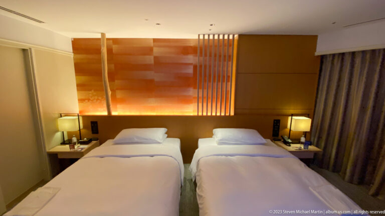 Twin hotel beds beneath a wood headboard with vertical slats and warm concealed lighting.