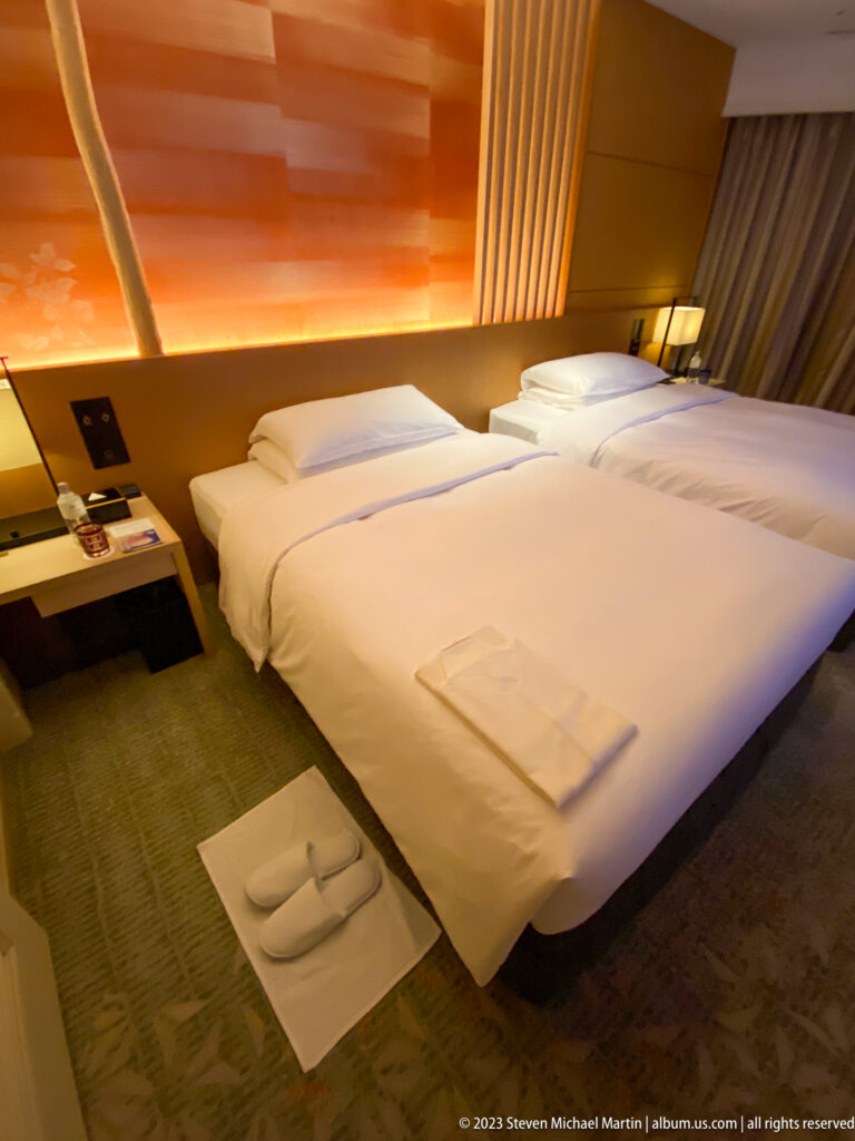 Diagonal view of two hotel beds with folded linens and amber-lit wood paneling.