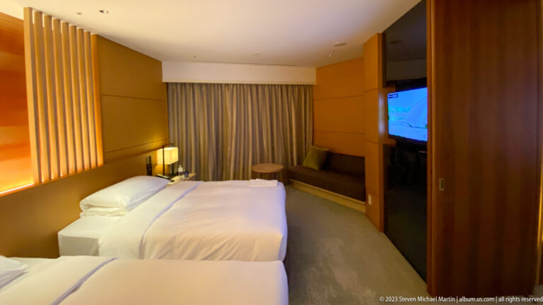 Twin hotel beds facing a television, curtained window, and small seating area.