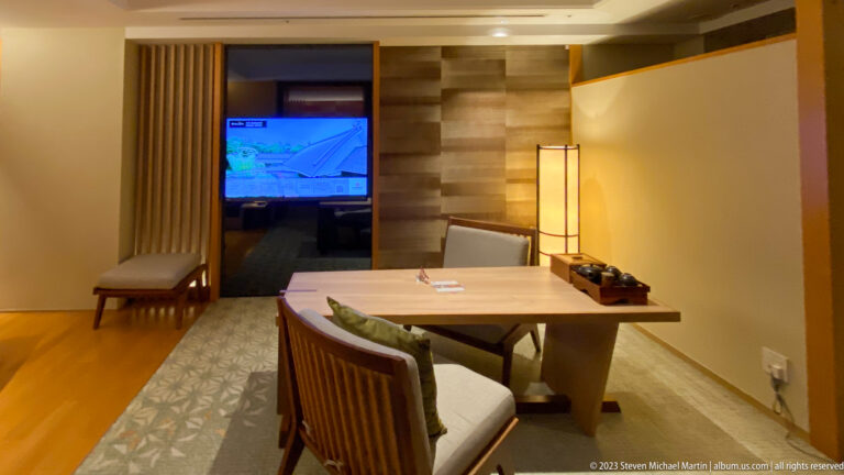 Wood table with upholstered chairs, television, bench, and floor lamp in a hotel suite.