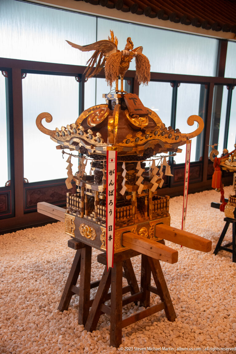 Gilded mikoshi with phoenix finial, ornate roof, paper streamers, and wooden carrying poles.