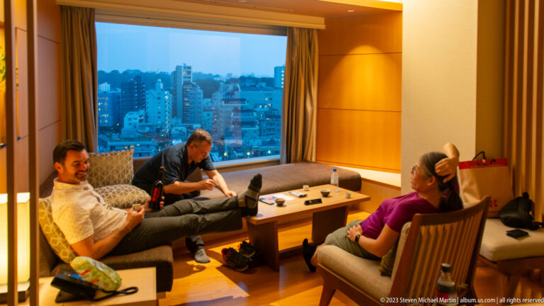 Three adults relaxing around a coffee table in a hotel suite with city view.