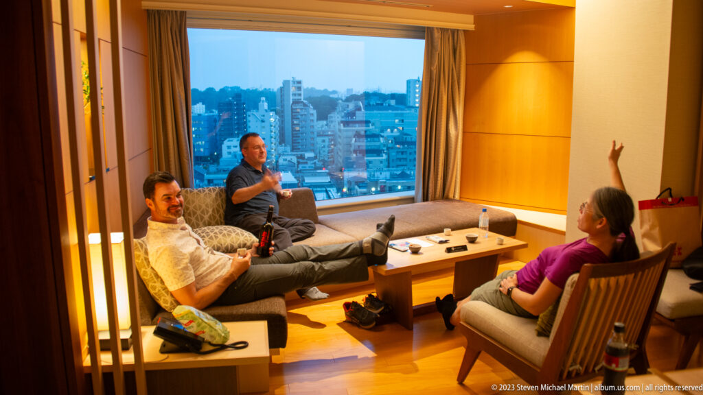 Three adults conversing in a warmly lit hotel suite beside a dusk city window.
