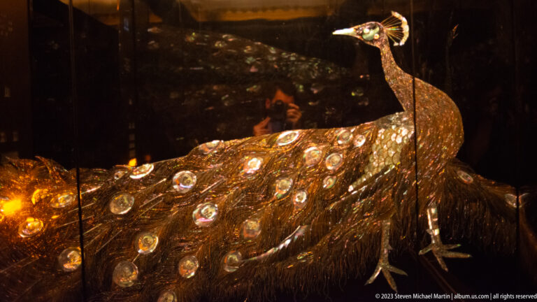 Side detail of a gilded peacock before an illuminated tail of reflective eye motifs.