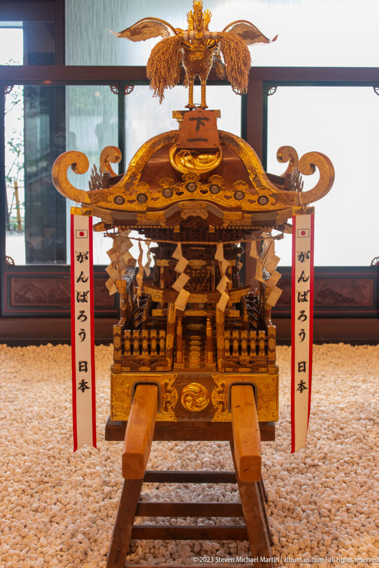 Frontal view of an ornate gilded mikoshi with phoenix crest, streamers, and projecting carrying poles.
