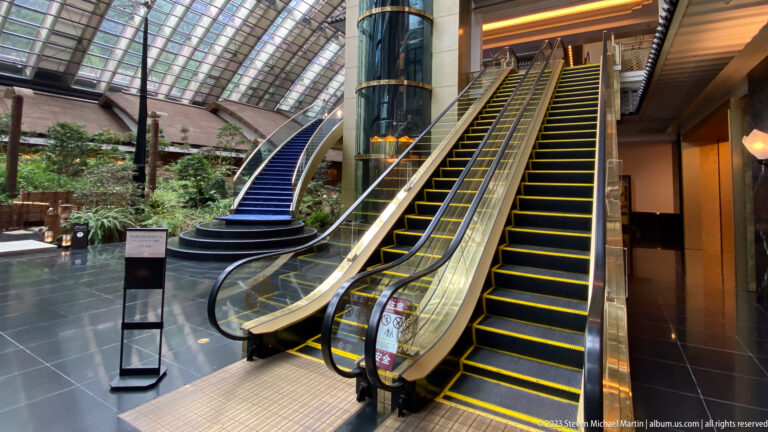 Two escalators beside a curved blue staircase and cylindrical glass elevator in a planted hotel atrium.