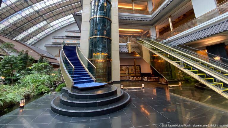 Wide hotel atrium with curved blue stair, glass elevator, escalator, balconies, and indoor plantings.