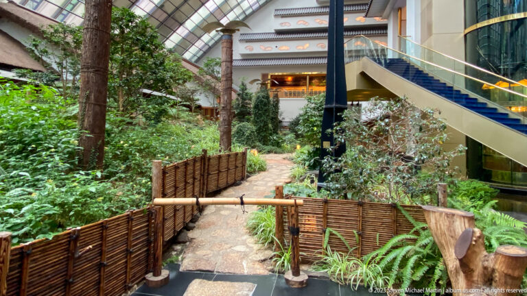 Stone path through dense indoor greenery beneath a staircase and balcony, edged with low woven fencing.