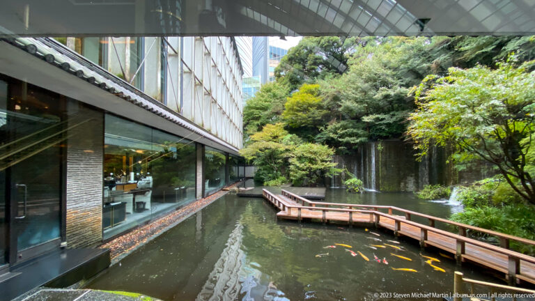 Curving wooden walkway beside a koi pond, glass-walled hotel interior, and dense Japanese garden planting.