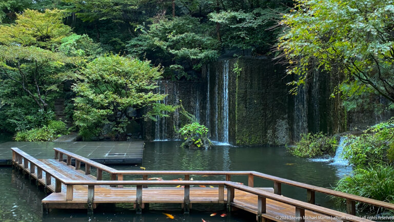Artificial waterfall spilling into a koi pond beside wooden garden railings and dense green foliage.