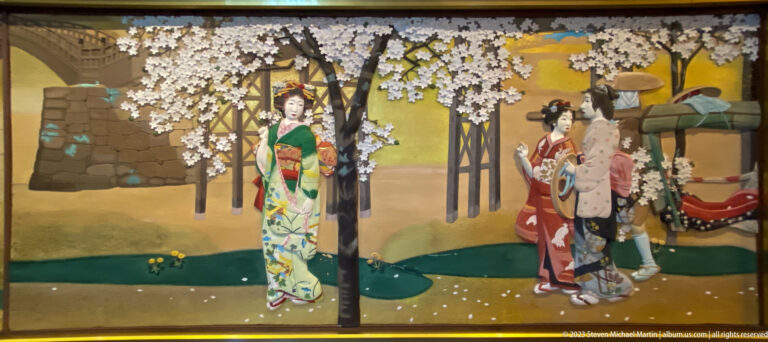 Women in colorful kimono gather beneath spring blossoms beside a stream and wheeled carriage.