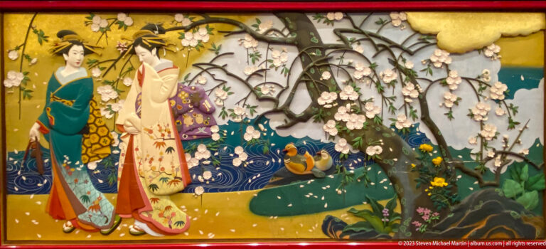 Three women in patterned kimono stand beside a stream with ducks, rocks, and flowering branches.