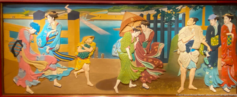 Women in colorful kimono walk with a child and umbrella-bearing attendant near a gate.
