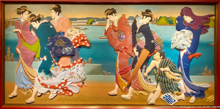 Women in patterned kimono assist a bending figure beside blue water and distant waterfront structures.