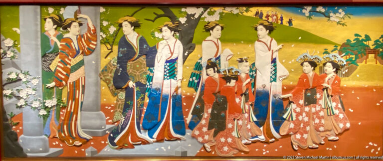 Adult women and girls in varied kimono gather beneath flowering branches in an open landscape.