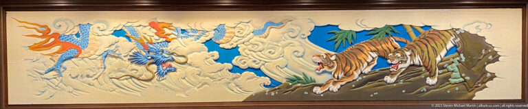 Dragon in clouds faces two tigers on rocky ground across stylized waves and bamboo.