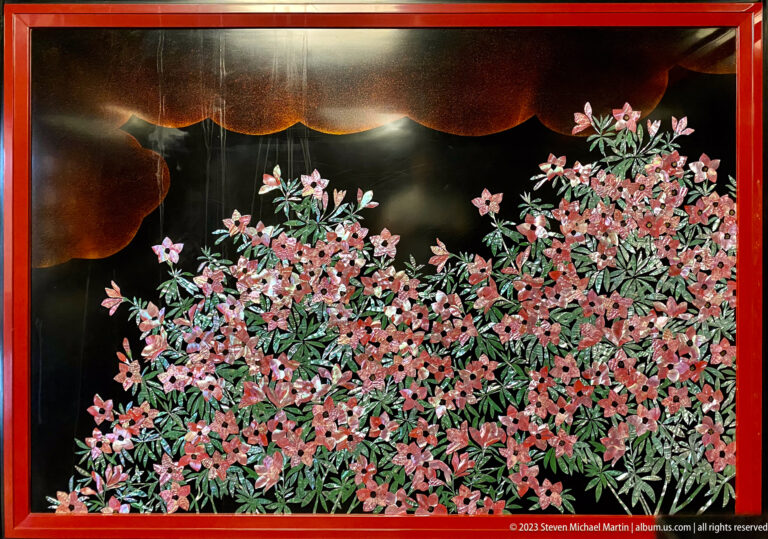Dense pink blossoms and narrow green leaves spread across a glossy black panel in a red frame.