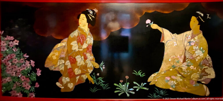 Two women in patterned kimono face each other beside dense pink blossoms on a glossy black ground.