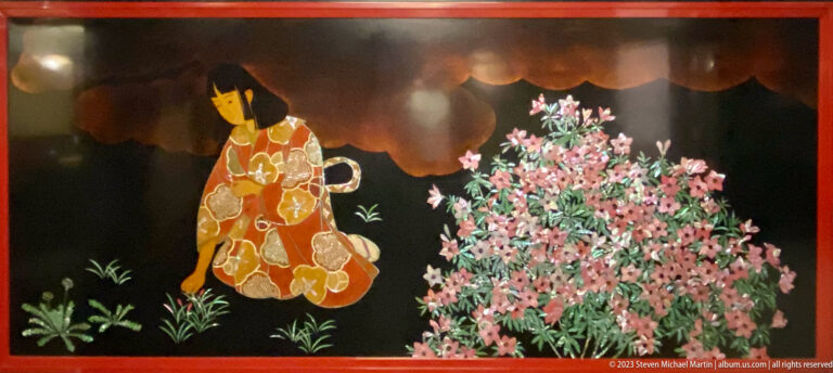 Solitary woman in a gold-patterned kimono stands beside dense pink blossoms on a glossy black panel.