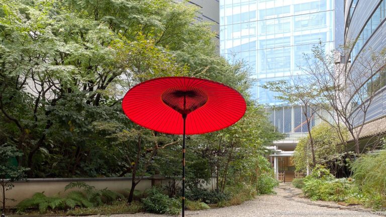Red Japanese-style parasol beside a gravel garden path surrounded by trees and modern hotel buildings.