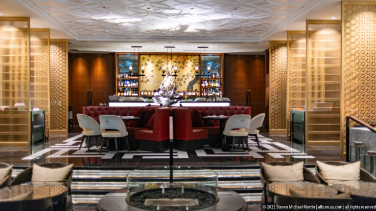 Symmetrical hotel lounge with red seating, pale lattice screens, and an illuminated central bar.