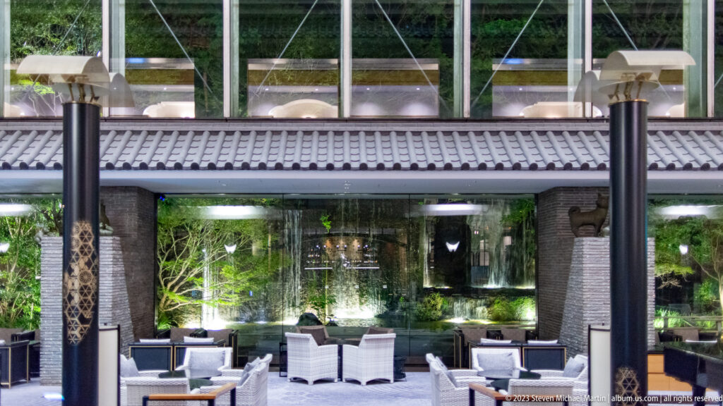 White lounge chairs beneath tiled eaves and dark columns inside a glass-covered courtyard.
