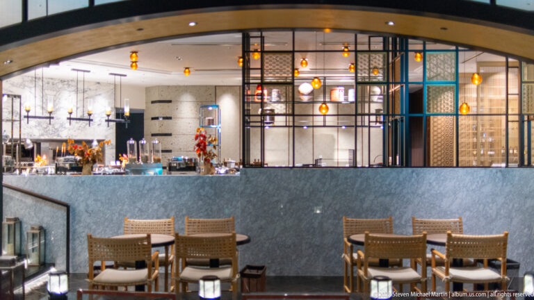 Dining tables and woven chairs before a stone partition, pendant lights, and a metal-framed restaurant interior.