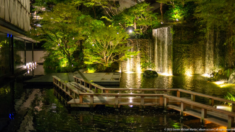 Illuminated waterfall, pond, trees, and wooden walkway in Hotel Gajoen’s garden at night.