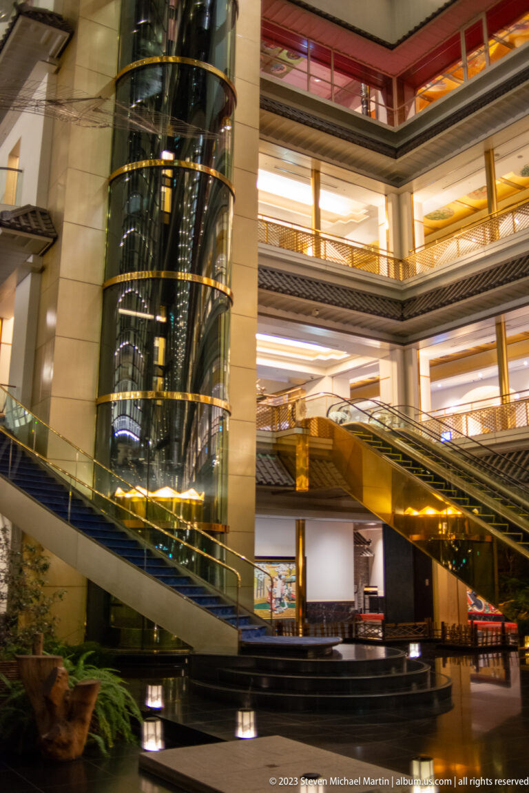 Multi-level hotel atrium with cylindrical glass elevator, escalators, balconies, and dark stone platforms.