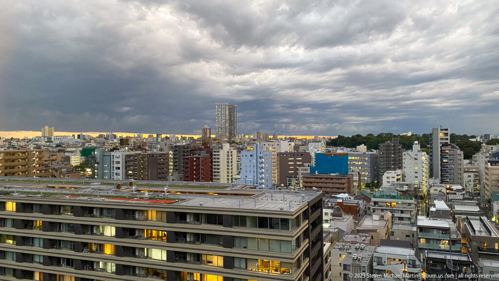 Wide Tokyo cityscape of apartment and commercial buildings beneath layered storm clouds.