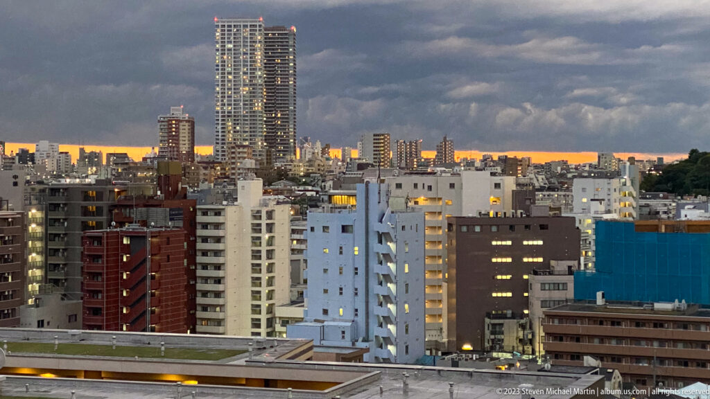 Tokyo high-rises at dusk beneath dark storm clouds and a narrow orange horizon.
