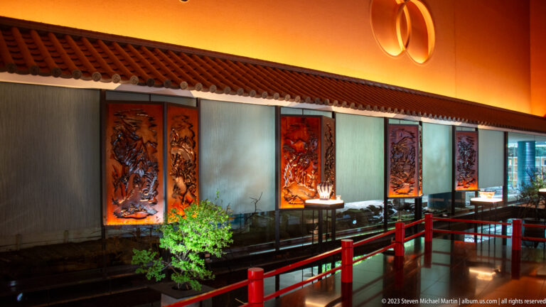 Interior gallery with carved panels, red railings, display lighting, and planted greenery.