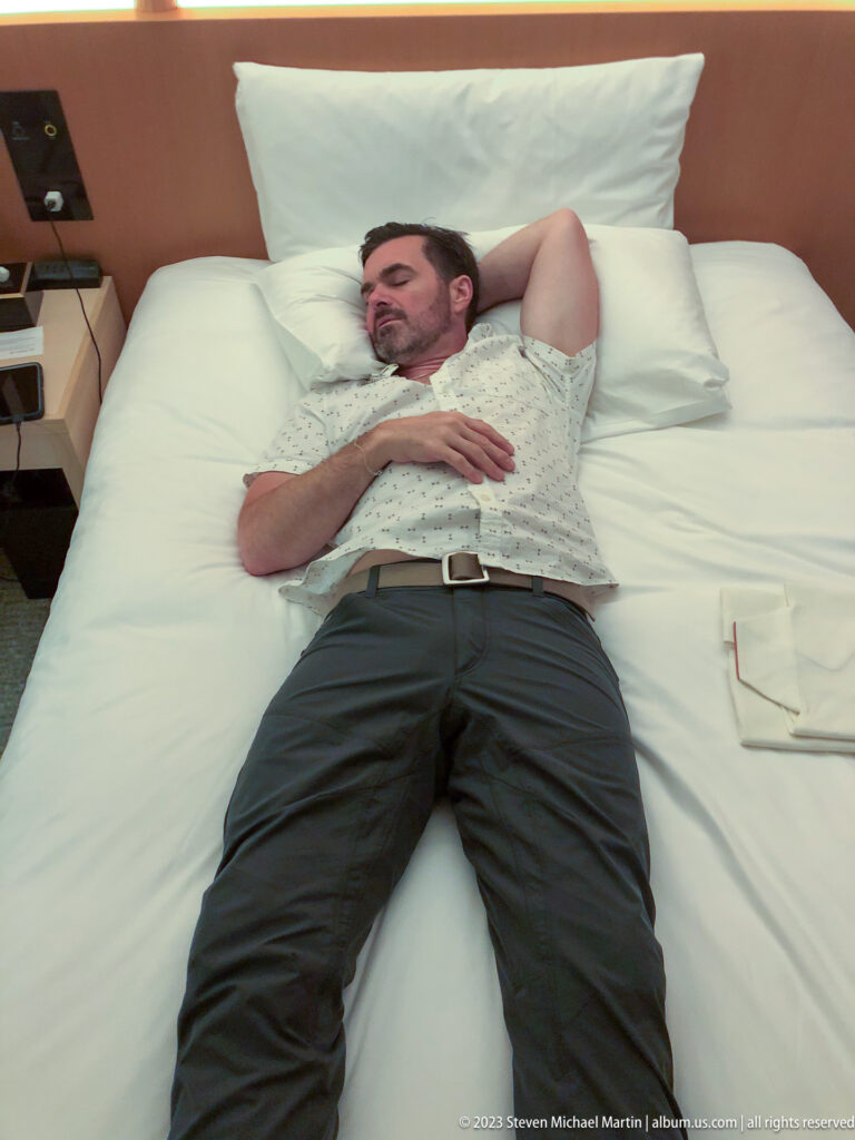 Male hotel guest reclining on white bedding with one arm behind his head.