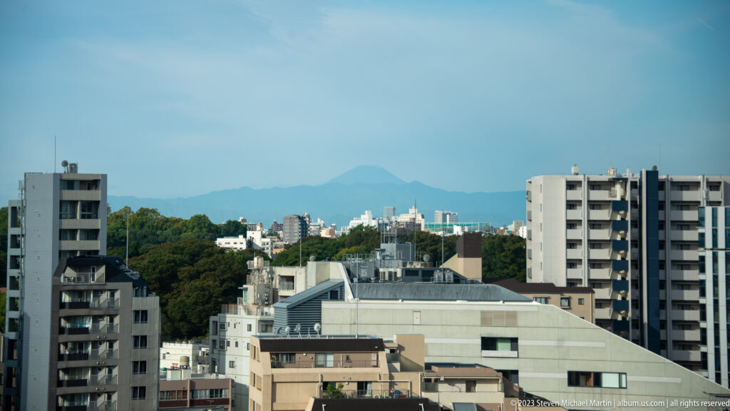 Faint Mount Fuji rising beyond Tokyo apartment buildings and a wooded ridge.