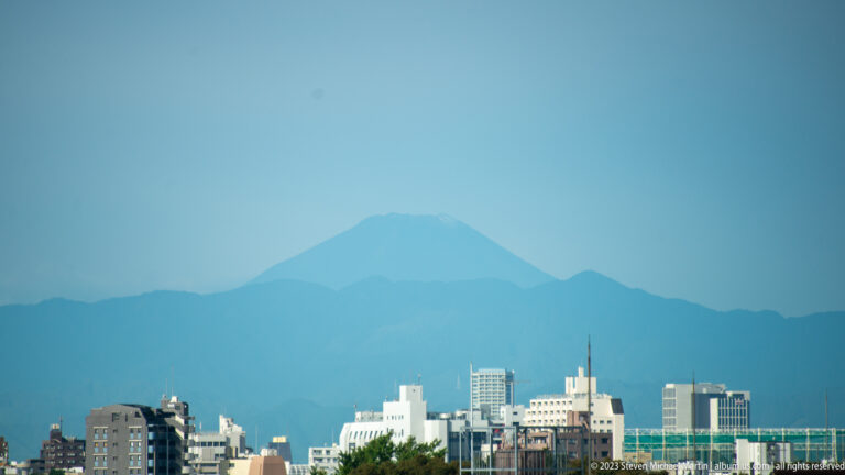Telephoto view of Mount Fuji rising above Tokyo rooftops and a darker foothill ridge.