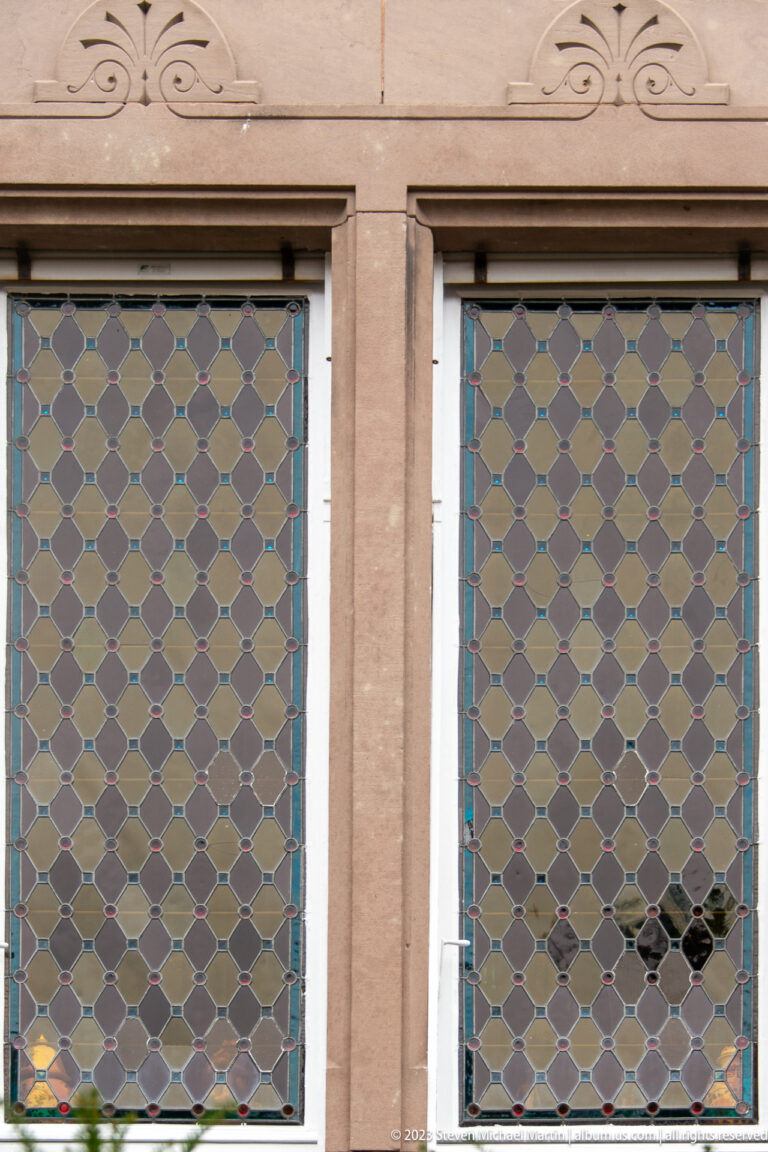 Two adjacent windows with diamond-patterned leaded glass in muted colors set in a pale façade.