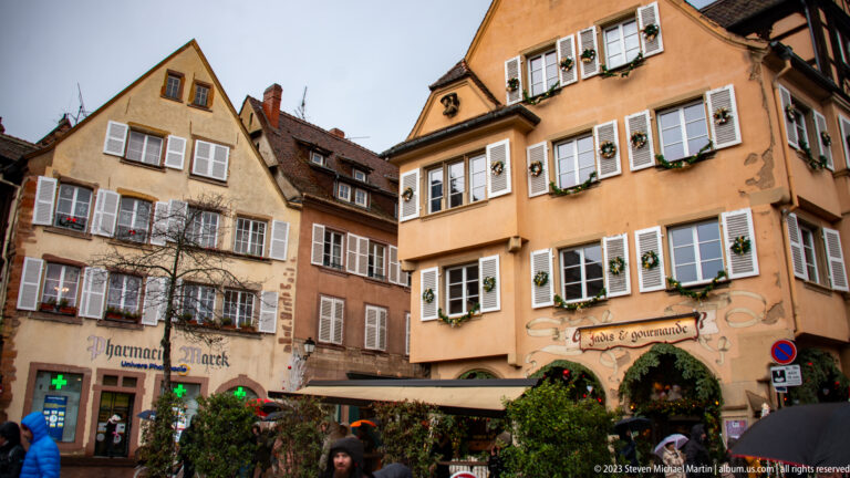 Warm-toned historic houses with rows of pale shutters and Christmas decorations above pedestrians in Colmar.