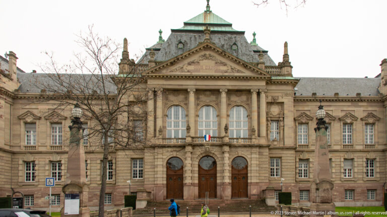 Formal stone civic façade with central columns, pediment, tall windows, green roof, and French flag above the entrance.