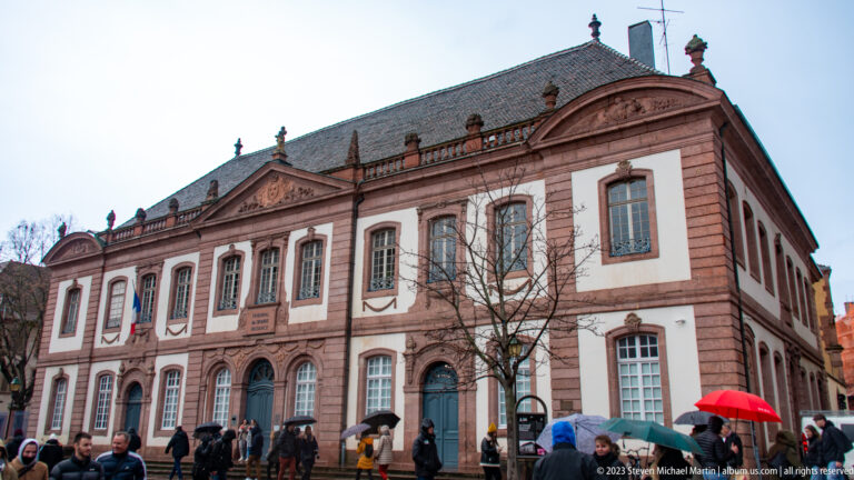 Large symmetrical public building with pink stone trim, arched lower windows, central pediment, and crowds with umbrellas.