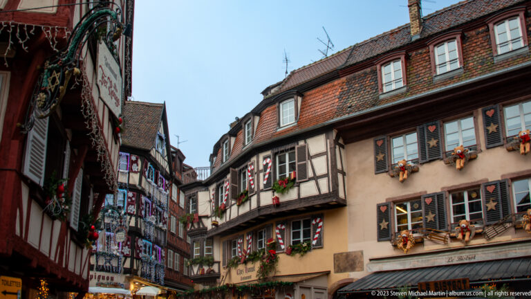 Narrow Colmar street lined with half-timbered houses, shutters, stars, greenery, and red-and-white Christmas decorations.