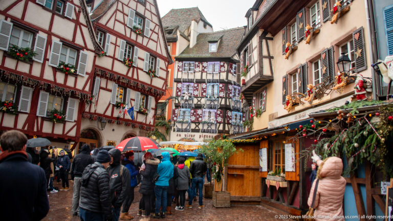 Crowds with umbrellas and raincoats move between Christmas stalls and colorful half-timbered houses.