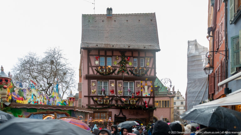 Narrow gabled half-timbered house with decorated windows rising above umbrellas and a Christmas market crowd.