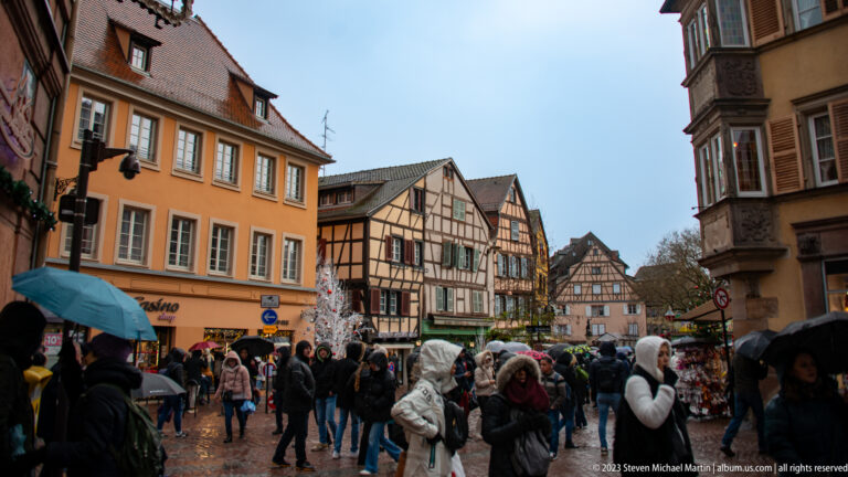 Pedestrians in winter clothing cross a wet Colmar street bordered by historic houses and Christmas decorations.