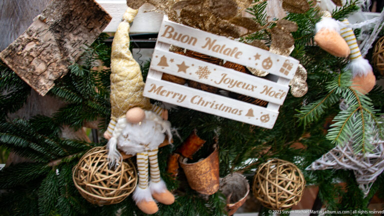 Wooden sign reading “Buon Natale,” “Joyeux Noël,” and “Merry Christmas” among evergreen branches and gnome ornaments.