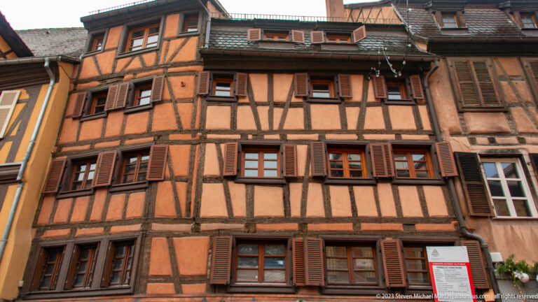 Close view of an ocher half-timbered façade with dark structural beams and rows of brown shutters.