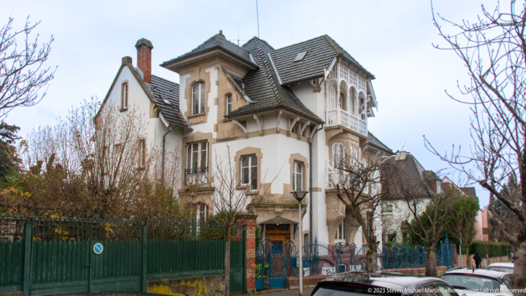 Asymmetrical historic villa with steep tiled roofs, projecting bays, balconies, and a green street fence.