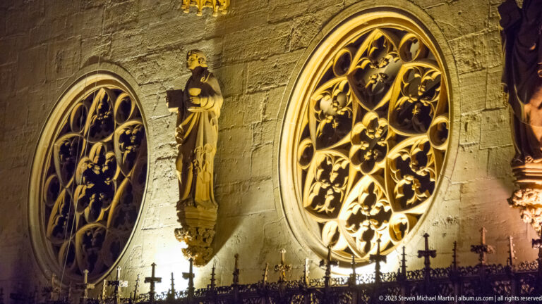 Two illuminated Gothic tracery windows flanking a small religious statue on a stone church wall.