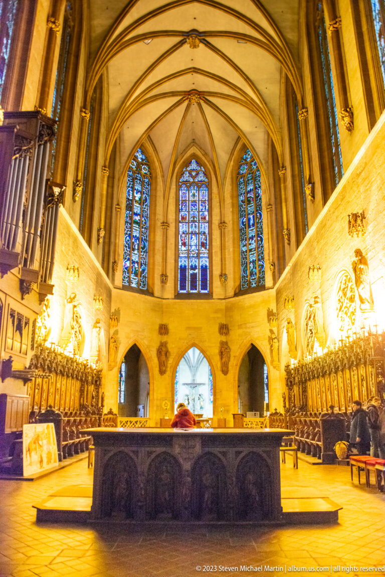 Gothic church choir with rib vaults, stained-glass windows, carved stalls, and a dark central liturgical platform.